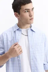 Camisa de popelina a cuadros, color celeste y blanco, de manga larga y corte holgado. Presenta un bolsillo frontal y cartera abotonada.