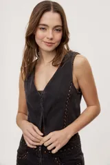 Chaleco corto de denim negro con escote en V y diseño sin mangas. Presenta terminación lavada y detalles laterales con entramado y tachas metálicas que aportan un toque distintivo.