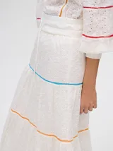 Falda larga blanca confeccionada en broderie de algodón con diseño de paneles escalonados, detalles de galones con transparencias en color azul y naranja, y pretina elástica con cordón ajustable.