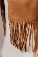 Chaleco de efecto gamuza en tono camel, sin mangas, con escote en V y diseño abierto. Presenta detalles de tachas metálicas, entramado artesanal en el frente y flecos largos en el ruedo.