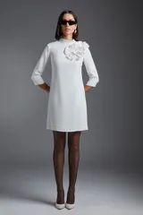 Vestido corto blanco de corte recto, con cuello alto, mangas 3/4 y aplique floral en el hombro.