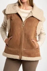 Chaleco estilo gamulán de corte oversize, confeccionado en material tipo gamuza color camel con interior y bordes de corderito sintético color crema. Presenta cierre frontal completo, bolsillos laterales y detalles de tiras decorativas en los hombros.