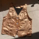 Chaleco sin mangas de color dorado con acabado brillante, escote en V y cierre frontal de tres botones. Presenta bolsillos ojal falsos en el frente y una pequeña etiqueta de la marca en el borde inferior.
