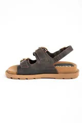Sandalias de cuero marrón oscuro, con doble tira ancha en el empeine y tira en el talón, todas con hebillas metálicas doradas. Cuenta con suela plana y gruesa de tono tostado con estriado inferior.