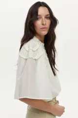 Blusa blanca con cuello mao, detalles de mini volados y bordados en el pecho y las mangas, con cierre frontal de botones y mangas largas abullonadas.
