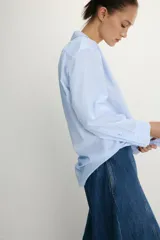 Camisa de popelín confeccionada en algodón, con cuello solapa, escote en pico y cierre frontal con botones. Presenta mangas largas con puños y un bajo redondeado asimétrico.