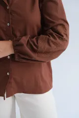 Camisa de color marrón chocolate, con cuello clásico, cierre frontal de botones y mangas largas abullonadas con detalle de tejido calado en los puños.