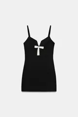 Vestido corto negro de escote en pico con tirantes finos y detalle de lazo blanco en el centro del pecho. Presenta un diseño ajustado con cierre de cremallera oculta en la espalda.