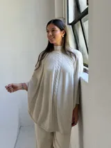Poncho de tejido de punto color beige con cuello alto y diseño de trenzas verticales en el frente. Presenta mangas integradas con puños acanalados y un corte holgado con ruedo redondeado.
