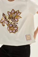 Remera blanca de algodón con cuello redondo y manga corta. Presenta un estampado bordado central con motivos florales y un animal (posiblemente un conejo o liebre) en tonos lila, morado y marrón.