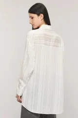 Camisa oversize de manga larga en color blanco con diseño de sutiles rayas verticales a tono y contratono, cuello camisero, bolsillo frontal en el pecho y cierre delantero con botones.
