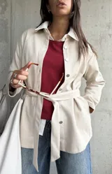Sobrecamisa de gamuza en color beige, con cierre frontal de botones metálicos, cuello camisero y lazo ajustable en la cintura. Presenta un diseño de corte recto y mangas largas.