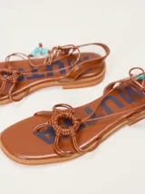 Sandalias planas de cuero color marrón, con diseño de punta cuadrada, tiras finas cruzadas y un detalle trenzado circular en el empeine. Cuentan con tiras para atar al tobillo decoradas con cuentas turquesas en las puntas.