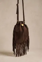 Cartera tipo bucket bag color marrón, confeccionada en material sintético con acabado gamuzado. Presenta un diseño con flecos largos en los laterales y base, cierre mediante cordón ajustable con detalle metálico circular y correa larga regulable.
