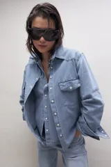 Camisa de jean celeste con cuello clásico, cierre frontal con botones a presión, dos bolsillos con solapa abotonada en el pecho y mangas largas con puños abotonados.