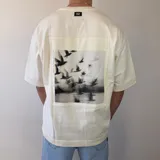 Remera oversize de algodón pesado en color blanco, con cuello redondo y moldería amplia. En la espalda cuenta con un gran parche rectangular estampado con una fotografía en blanco y negro de aves volando sobre el agua y texto tipográfico al margen.