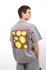 Remera de algodón con efecto lavado en color gris, de corte holgado y manga corta. Presenta un estampado cuadrado en la espalda con fondo marrón y círculos amarillos, junto con la inscripción CAPER.
