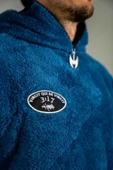 Canguro de textura tipo peluche en color azul, con cierre frontal parcial, cuello alto y bolsillo tipo canguro. Presenta un parche ovalado en el pecho con diseño gráfico.