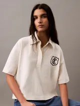 Chomba color crema de punto de algodón con cuello clásico, tapeta de botones oculta y mangas cortas. Presenta un corte holgado y un escudo bordado con el logo de Calvin Klein en el pecho.