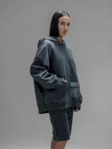 Hoodie oversize de color gris oscuro, confeccionado en frisa de algodón con recortes en textiles técnicos. Presenta capucha forrada, bolsillo frontal tipo canguro y un bolsillo adicional con solapa en la parte inferior.