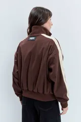 Cazadora de estilo deportivo en color marrón, confeccionada en tejido tipo gamuza. Presenta cierre frontal completo, cuello alto, puños y ruedo acanalados, y franjas laterales en tono rosado. Incluye el logo de la marca bordado en el pecho.