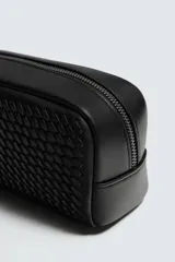 Neceser rectangular negro con textura trenzada en todo su exterior. Posee un compartimento principal con doble cierre de cremallera y un bolsillo interior con cierre. Incluye un asa de mano lateral.