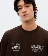 Remera de algodón color marrón con cuello redondo y mangas cortas. Presenta un diseño gráfico de estilo barroco con una figura alada y la inscripción 'Humble' en la espalda.