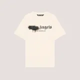 Camiseta de manga corta color beige con cuello redondo. Presenta un estampado frontal con el logo de Palm Angels en tipografía gótica negra, con efecto de pintura en aerosol y goteo, y el texto "LOS ANGELES" debajo.
