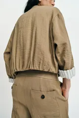 Chaqueta corta de color verde militar, con cuello solapa, bolsillos delanteros con solapa y cierre frontal de botones ocultos. Presenta mangas amplias con vuelta en los puños que muestran un forro a rayas.