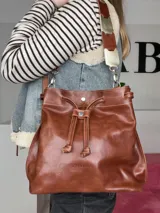 Bolso tipo bucket Chabas modelo Marin, confeccionado en cuero genuino de color marrón. Cuenta con diseño de cordón ajustable fruncido en la parte superior, manija corta decorativa de cuero con detalles metalizados y una correa larga intercambiable.