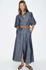 Vestido midi de denim azul con cuello camisero, mangas abullonadas y cierre frontal oculto con botones. Incluye un cinturón marrón con hebilla metálica.