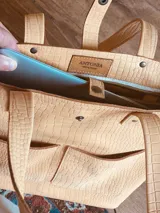 Bolso tipo tote de cuero sintético color beige con textura tipo cocodrilo. Tiene doble asa de mano y correa larga extraíble y ajustable. Cuenta con cierre principal, dos bolsillos externos y un bolsillo interno con cierre.