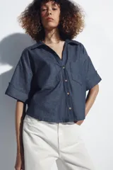 Camisa crop color blanco confeccionada en mezcla de lino, con cuello solapa, escote en pico, mangas cortas con trabilla, bolsillos de plastrón en el pecho y cierre frontal con botones.