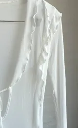 Vestido largo de microtul blanco, con escote en V profundo y mangas largas acampanadas. Presenta aberturas laterales en la falda y cierre con lazos en el frente. Es una prenda transparente.