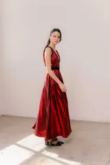 Vestido largo rojo brillante con escote en V profundo, espalda abierta baja y breteles anchos. Presenta detalles en terciopelo negro que enmarcan el escote, recorren el frente y la espalda, y una faja doble en la cintura ajustable con cinto y hebillas. La falda tiene un corte frontal que permite ver sutilmente la pierna.