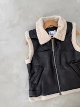 Chaleco de cuero sintético negro con interior y bordes de corderito sintético color crema. Presenta cierre frontal metálico, bolsillos laterales y correas ajustables con hebilla en el ruedo.