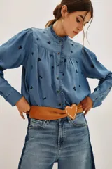 Camisa de lyocell color azul con estampado floral pequeño en tono oscuro. Presenta cuello alto con volados, cierre frontal con botones y mangas largas abullonadas con puños abotonados.