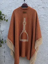 Ruana de tejido grueso con diseño de espiga en color tostado, con bordado central de un gaucho a caballo y la marca Bagual. Presenta terminaciones con flecos largos en color crema en todo su borde.