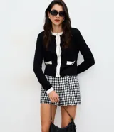 Cardigan corto de punto negro con detalles en blanco en la botonera, cuello redondo y bolsillos frontales. Cuenta con cierre frontal de botones perlados y mangas largas.