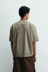 Camiseta de corte relajado con cuello redondo y manga corta. Presenta un efecto de lavado desgastado en tonos marrones y un estampado frontal de una cabeza de alienígena en color verde vibrante.