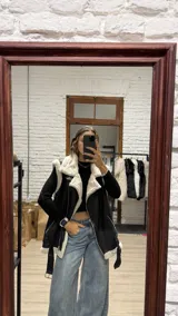 Chaleco estilo gamulán de cuero sintético negro con interior y bordes de corderito sintético color crema. Presenta cierre frontal, bolsillos laterales con cierre y cinturón ajustable en el ruedo.