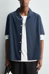 Camisa de manga corta con cuello bowling, confeccionada en popelín de algodón. Presenta un corte relajado, cierre frontal con botones y aberturas laterales en el bajo.
