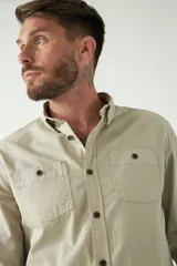Camisa de hombre color beige con efecto stone wash, corte regular, cuello clásico, cierre frontal con botones, dos bolsillos con solapa abotonada en el pecho y mangas largas con puños abotonados.