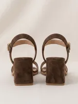 Sandalias de gamuza color marrón chocolate, de taco cuadrado medio y punta cuadrada. Tienen una tira delantera ancha y una tira ajustable al tobillo con hebilla metálica dorada.