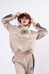 Canguro oversize color beige con capucha, confeccionado en frisa. Presenta tres rayas negras laterales en las mangas, bolsillo frontal tipo canguro y estampado gráfico con el logo de la marca en el pecho.