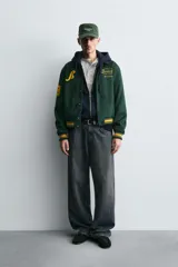 Cazadora tipo varsity de corte relajado confeccionada en tejido efecto ante color verde oscuro. Presenta cuello solapa, cierre frontal con botones a presión y puños y bajo acanalados con rayas en contraste. Incluye parches bordados en color amarillo en el frente y mangas.