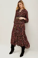 Vestido midi de corte holgado con estampado ikat en tonos bordó y tierra. Presenta cuello redondo con lazos, mangas largas abullonadas con puños elásticos y falda con volados escalonados. Incluye un cinturón ancho de estilo rústico con tachas y flecos.