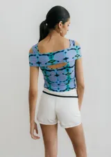 Top corto de jersey con estampado abstracto floral en tonos pastel (blanco, lila, verde, marrón). Presenta un diseño cruzado asimétrico en el busto que crea un escote tipo *off-shoulder* en un lado y manga corta en el otro.