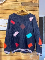Buzo de lana merino color azul marino, tejido a mano con la técnica de dos agujas, con cuello redondo y diseño holgado. Presenta apliques de retazos de cuero de diferentes formas geométricas y colores (rojo, celeste, rosado, bordó, naranja) cosidos a mano. Los puños, el dobladillo y el cuello cuentan con detalles de rayas en tonos rosado, celeste y azul.