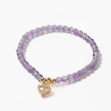 Pulsera elástica compuesta por cuentas de piedra amatista en tono lila, con un dije central de corazón calado con incrustaciones de cristales y detalles en metal dorado.